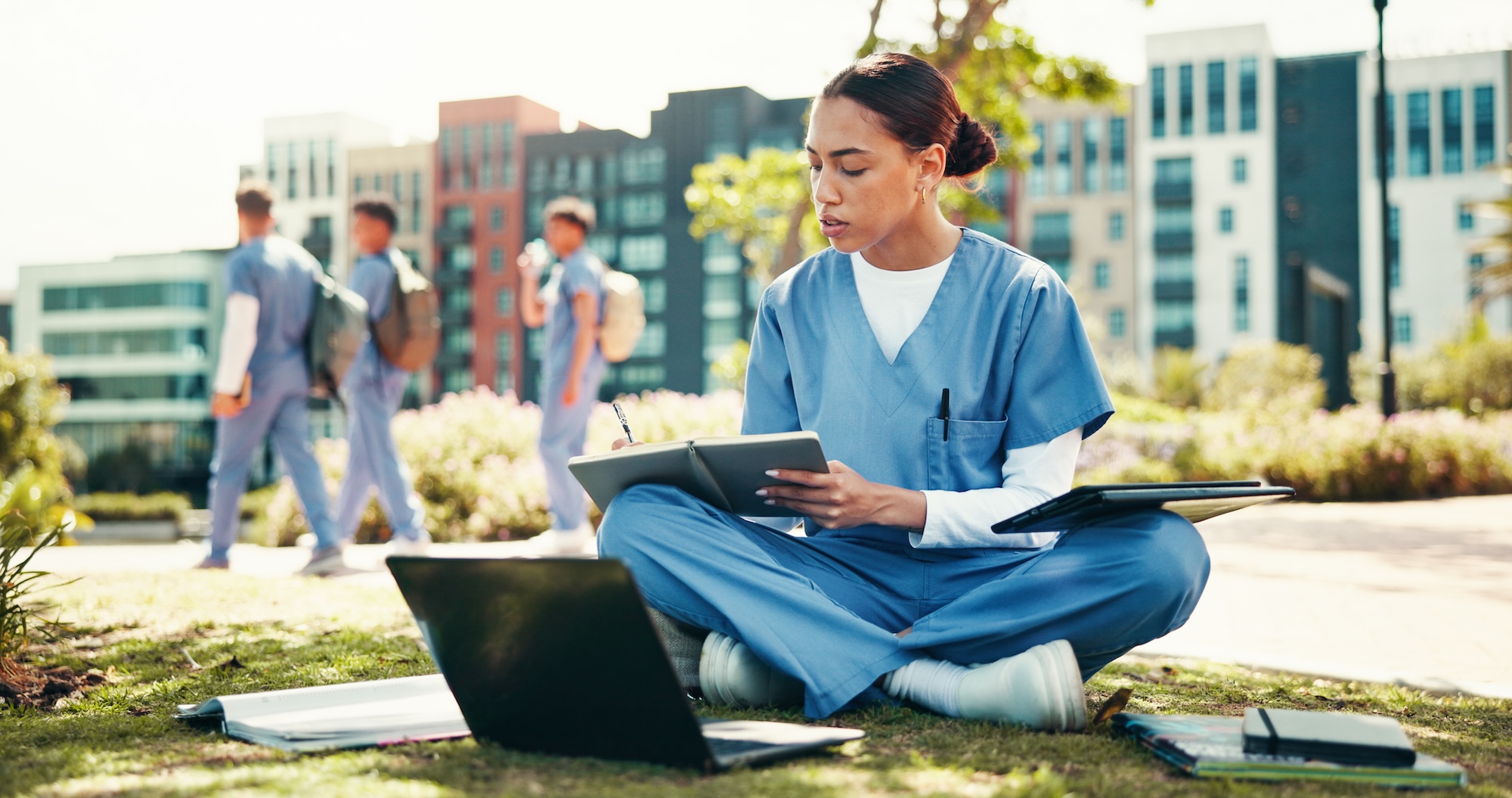 A woman in blue scrubs sitting on the grass with a laptop and a notebook