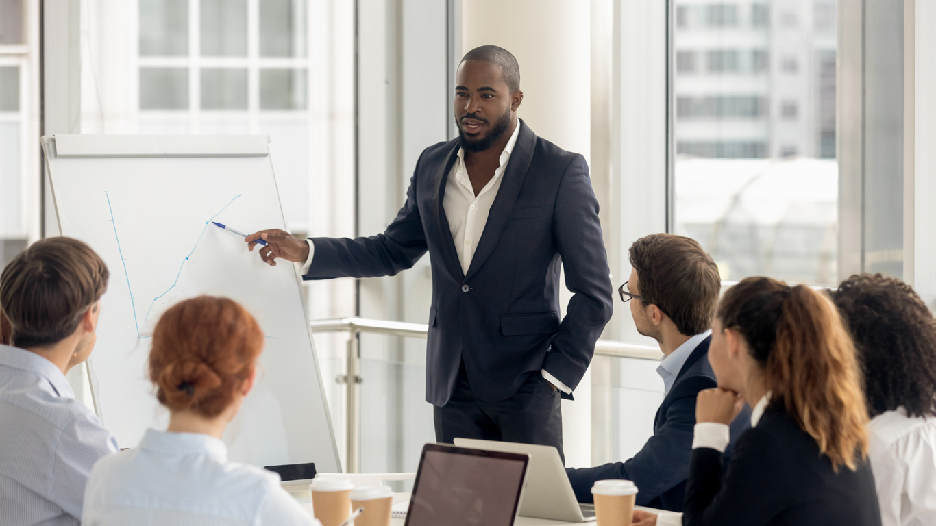 A man in a suit pointing at a whiteboard with other people sitting around him