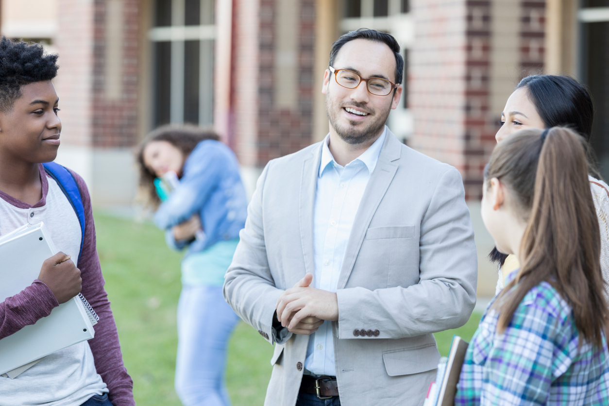 Confident high school teacher or principal talks with a group of high school students outdoors on campus.