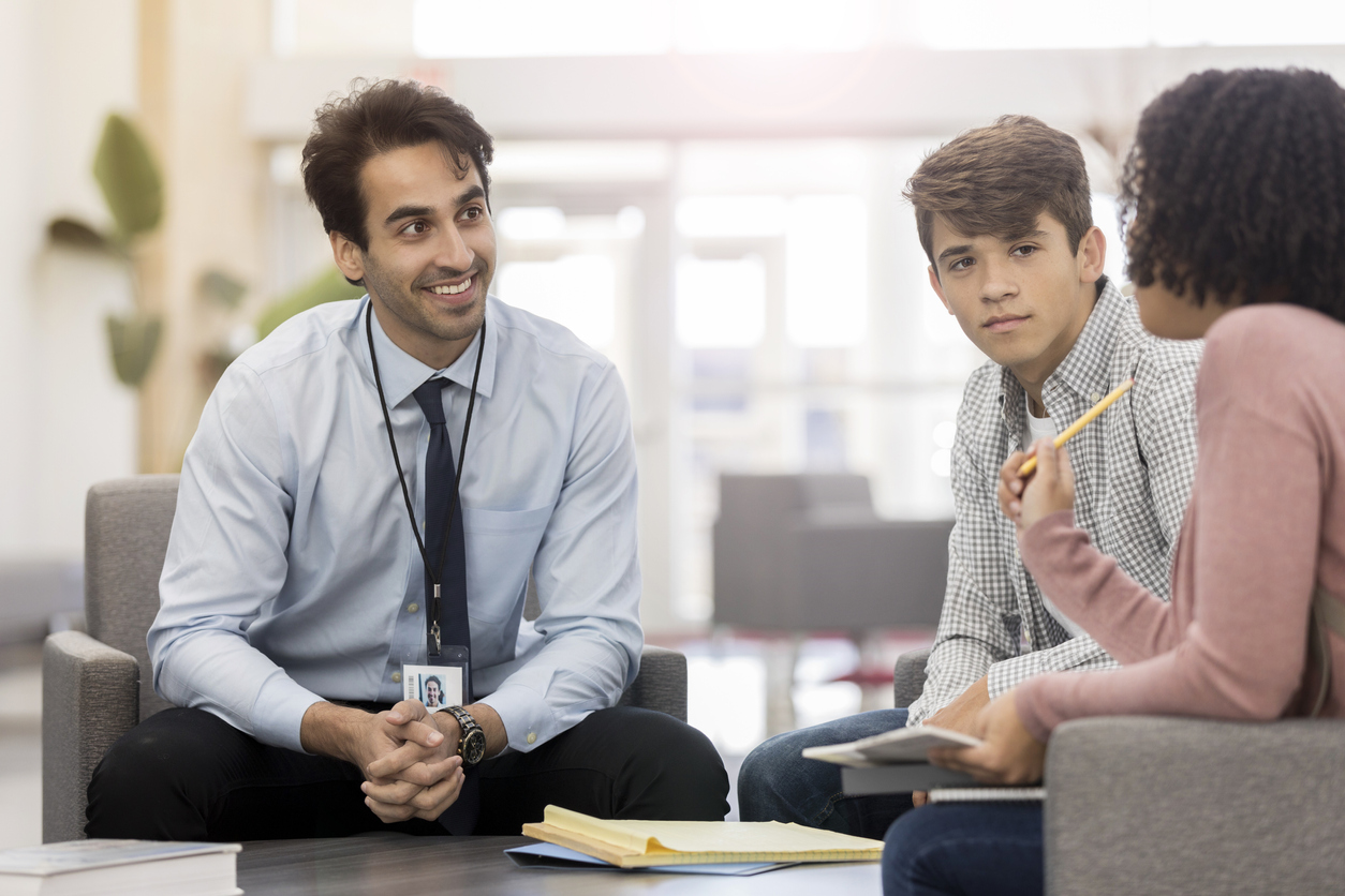 A cheerful high school principal sits around a coffee table in his high school lobby with two students. He listens attentively as an unrecognizable student shares a concern.